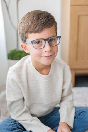 Child wearing glasses and cream sweater poses casually on floor.