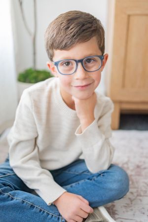 Child in glasses and cream sweater poses playfully on floor.