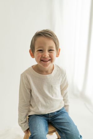Child in white sweater and jeans sitting against bright white background.