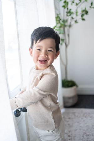 Young child in beige sweater playfully poses against white wall with indoor plants.