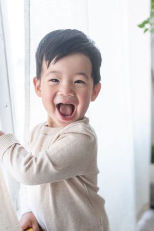Happy toddler in cream colored sweater playfully hiding their face while smiling.