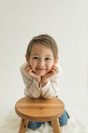 Cheerful child rests head on hands while leaning on a wooden stool against white background.