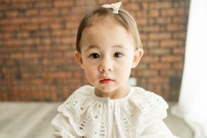 Small child in white lace dress poses against brick wall background.
