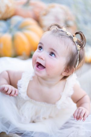 Baby in white dress smiles joyfully with pumpkins in the background.