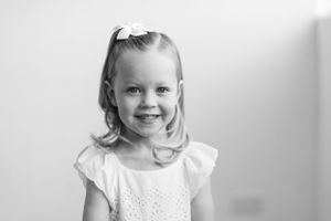 Black and white portrait of joyful child wearing white dress and bow in light colored hair.
