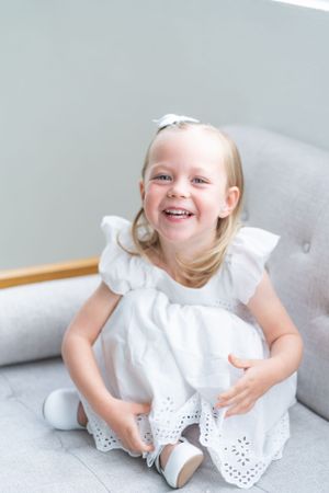 Young child in white eyelet dress sitting and laughing on gray sofa.