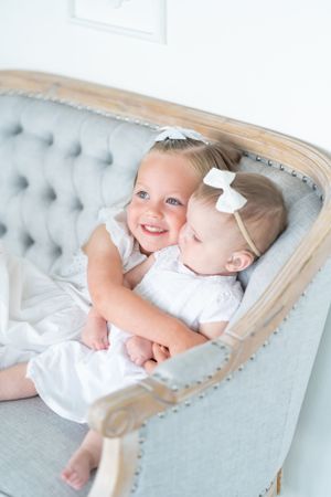 Two children in matching white outfits cuddling on a gray tufted settee.
