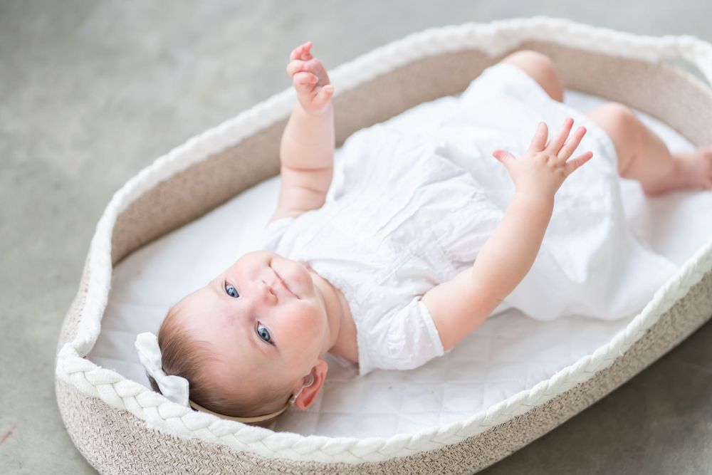 Happy baby in white outfit lying and playing in cream-colored bassinet.