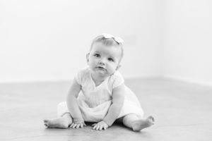 Black and white photo of baby in white dress sitting on floor.