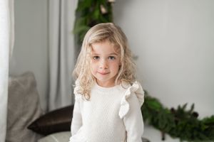 Young child in white knit sweater poses against grey wall with indoor plants.