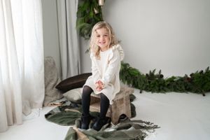 Child in white sweater sits on decorative blanket near window with greenery.