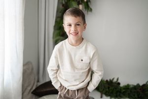 Young child in cream colored sweater stands by window with indoor plants.