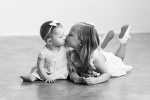 Black and white image of two children in white dresses sharing affectionate moment.
