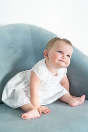 Baby in white dress sitting on light blue chair with upward gaze.