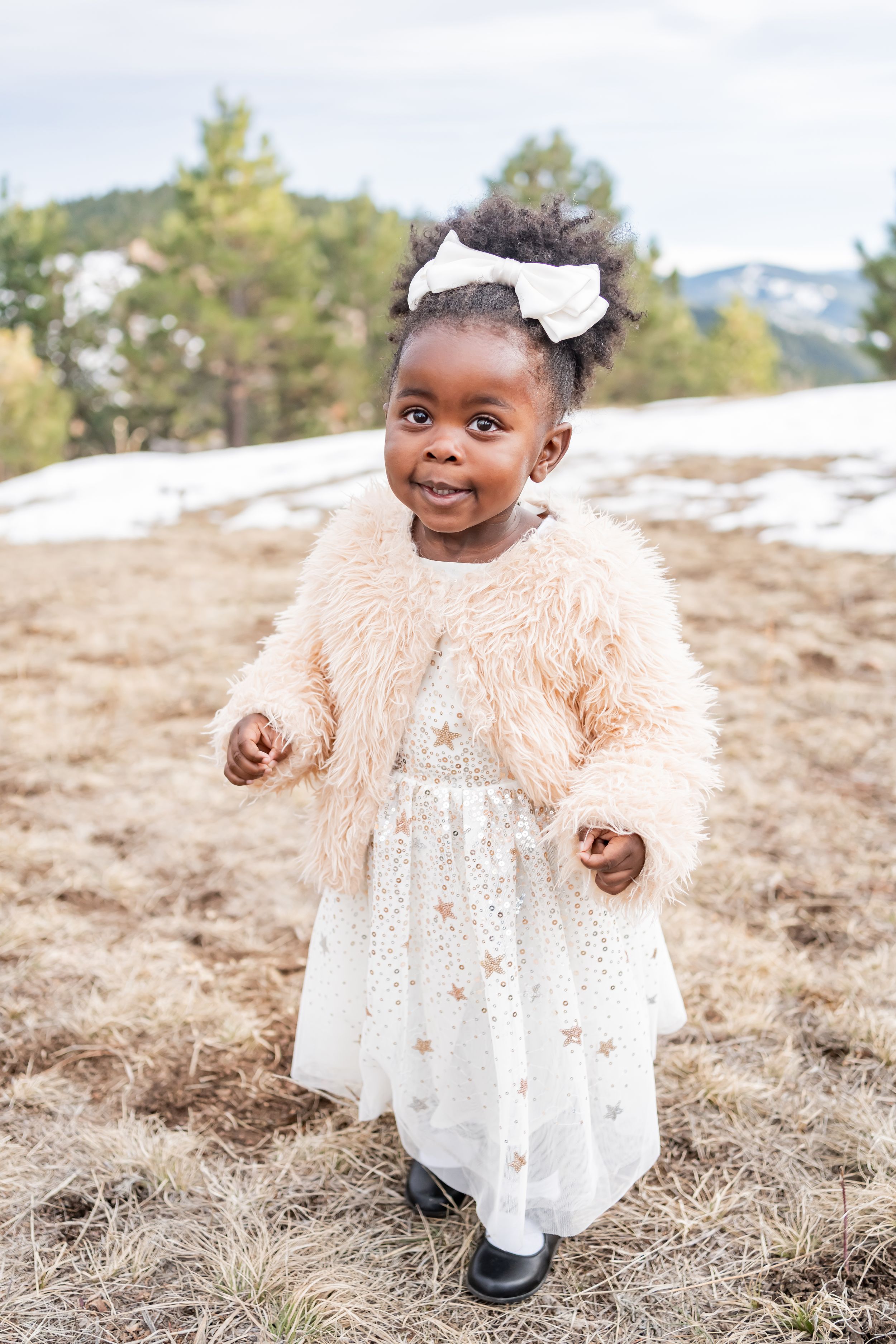 Child in fuzzy peach coat and white dress smiling outdoors in snowy landscape.