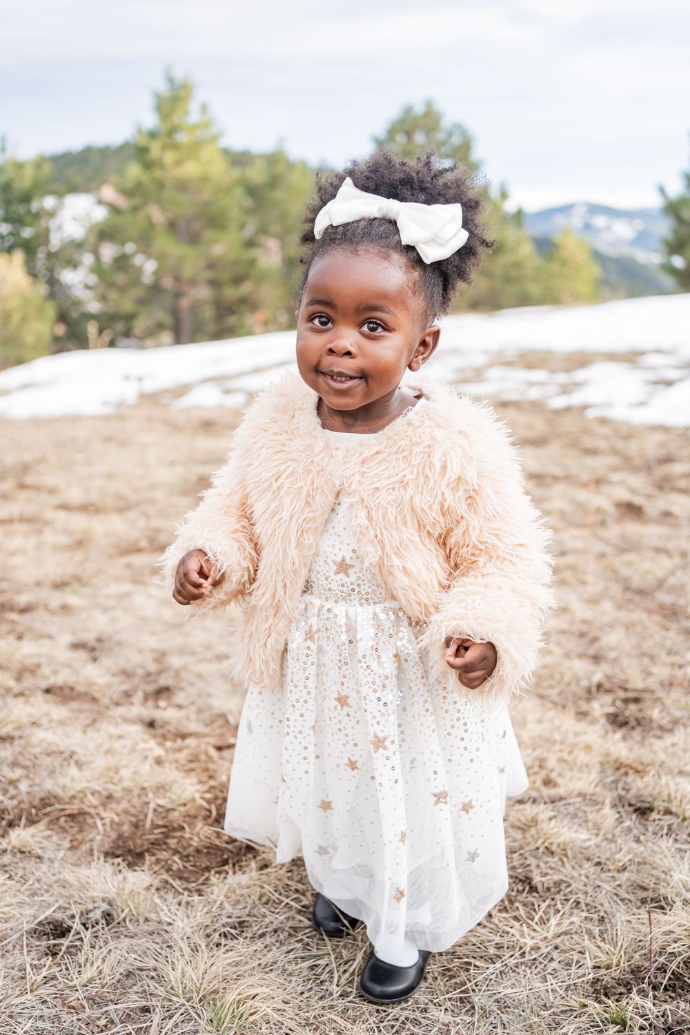 Child in fuzzy peach coat and white dress smiling outdoors in snowy landscape.