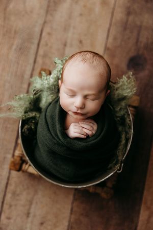 Sleeping newborn nestled in dark green wrap in vintage metal bowl against wooden background.