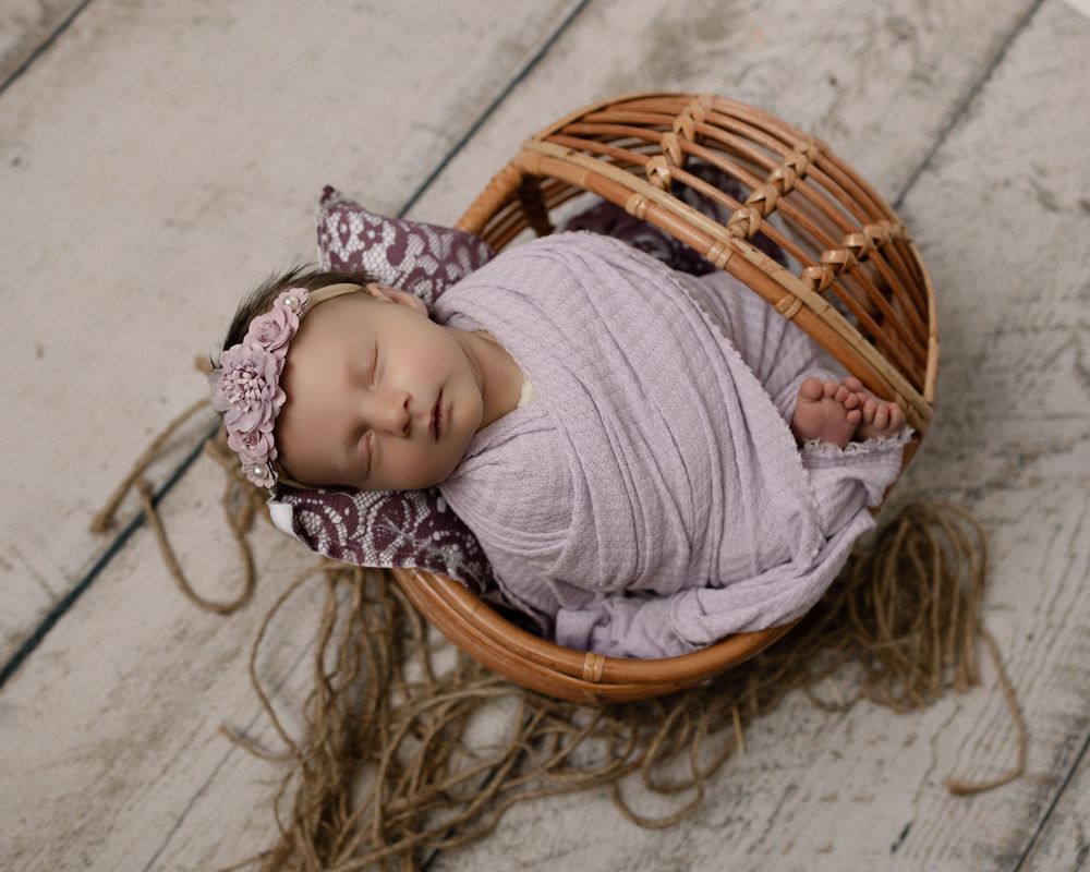 Newborn baby girl swaddled in lavender wrap with floral headband, sleeping peacefully in a wicker basket.