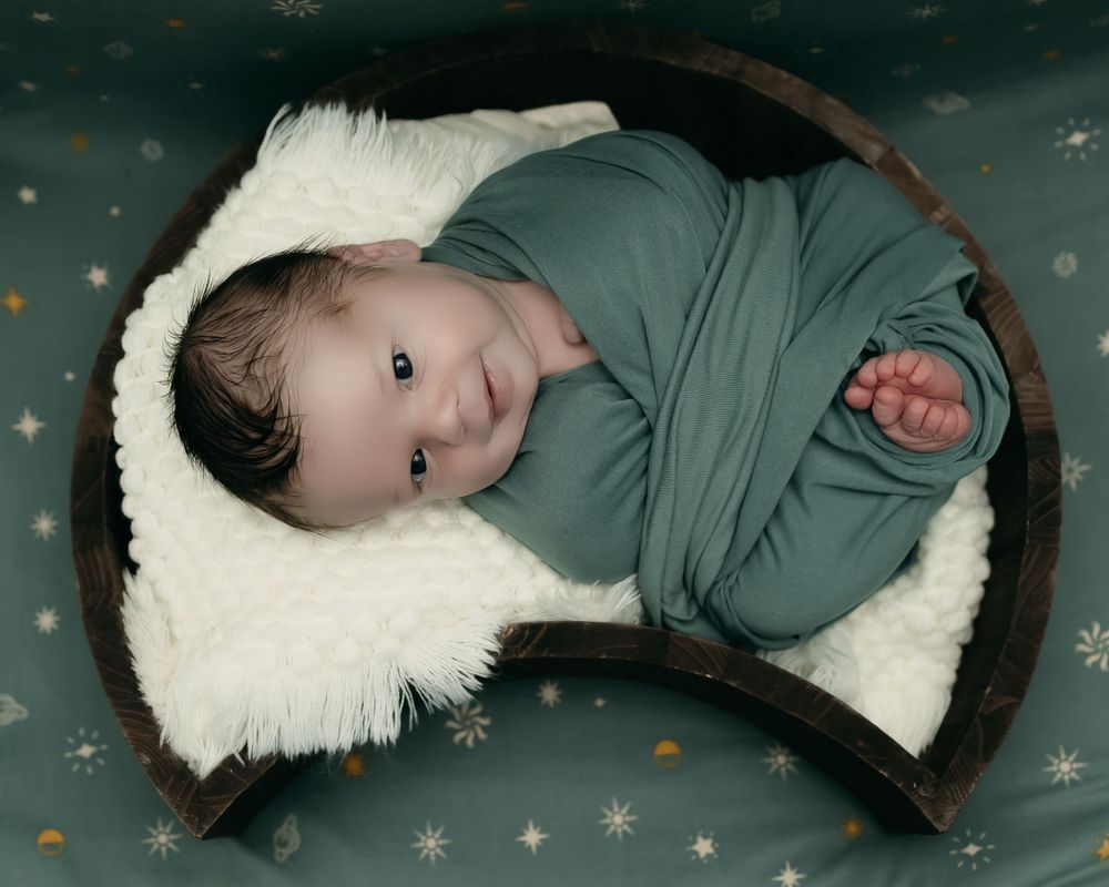 A baby wrapped in sage green cloth rests on a cream-colored blanket with stars in the background.