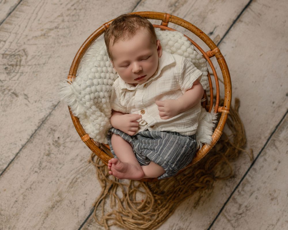 Newborn baby sleeping peacefully in a round wicker basket with cream knit blanket, wearing white shirt and striped shorts.