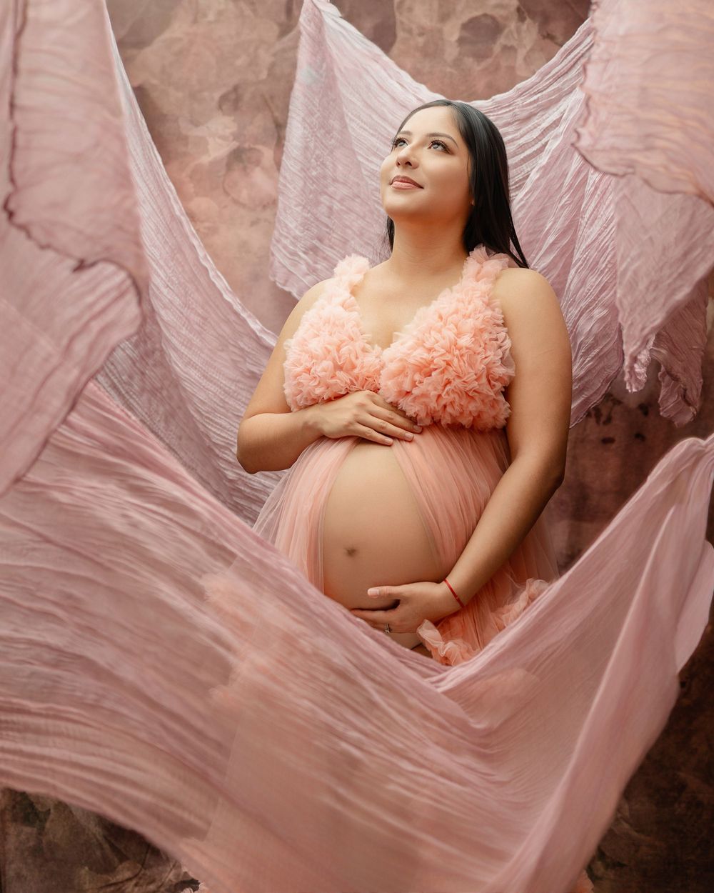 Pregnant woman in flowing pink ruffle maternity gown with fabric wings against marble backdrop.