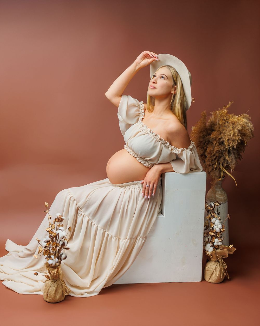 Pregnant woman in cream off-shoulder dress and white hat sits elegantly beside dried floral arrangements on terracotta background.