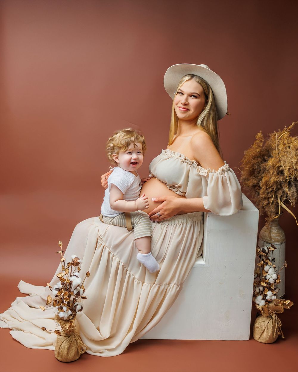 Pregnant woman in cream off-shoulder dress and hat poses with toddler boy in studio maternity photo.