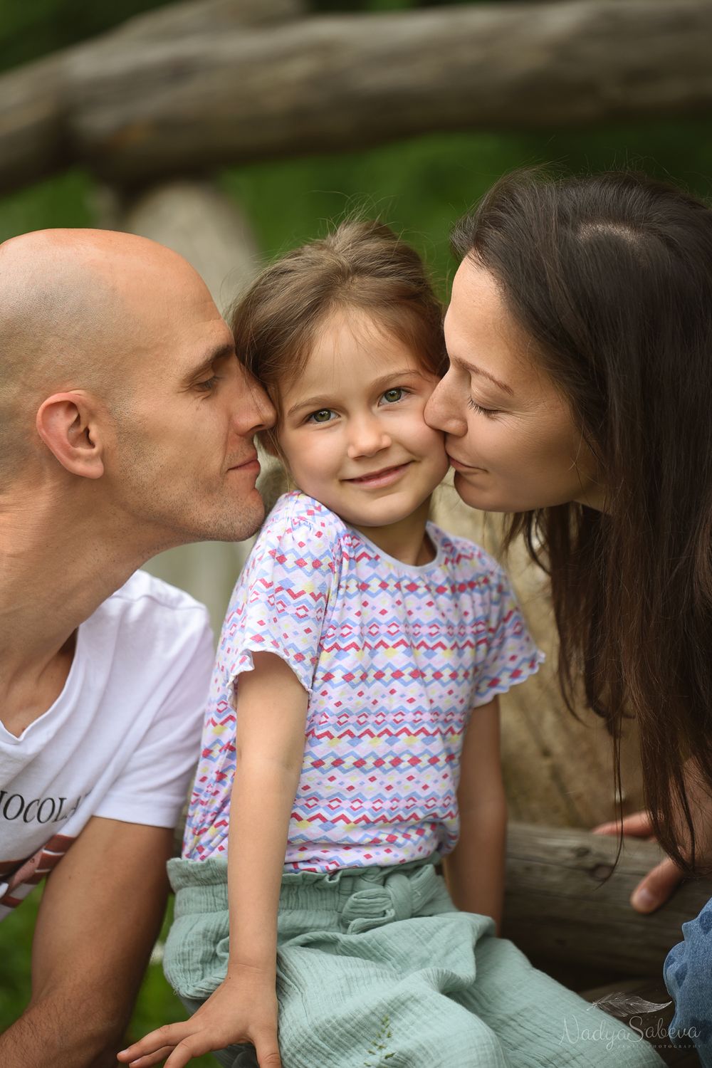 A loving family moment outdoors as parents give their young child sweet kisses from both sides.