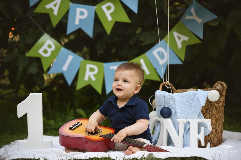 Smiling baby at first birthday outdoor photoshoot with guitar and blue banner decorations.
