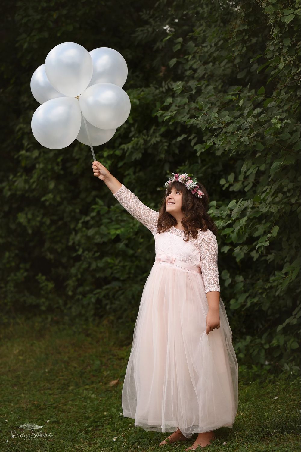 Dreamy outdoor scene with a figure in pink tulle dress and floral crown holding white balloons against green foliage.