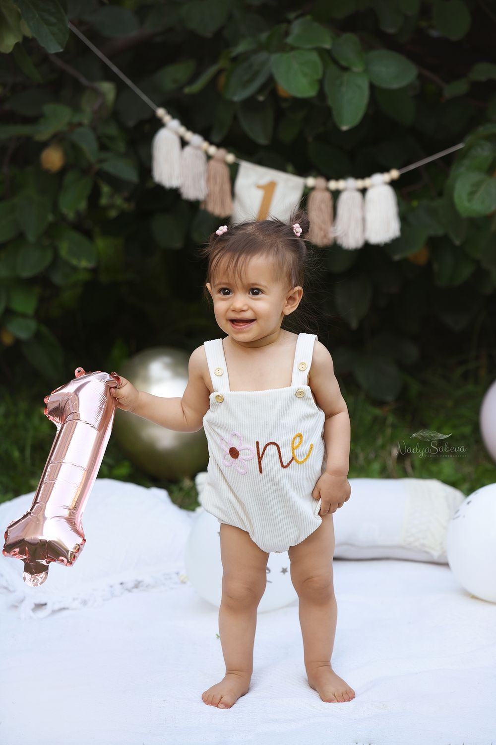 A small child in a white romper holds a pink number one balloon against a green leafy backdrop with party decorations.