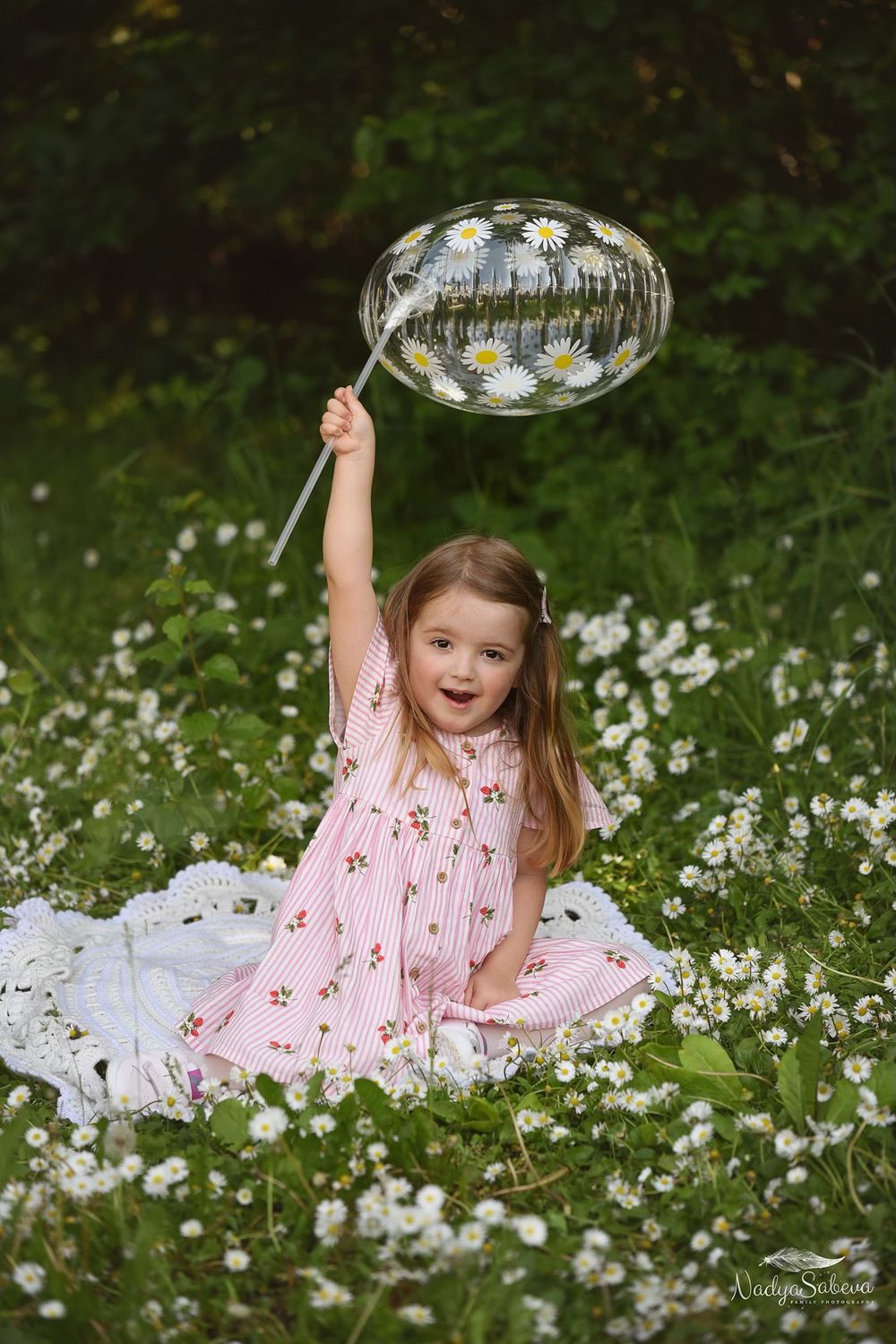 A child in a pink dress sits among daisies while holding up a large bubble against a blurred green background.