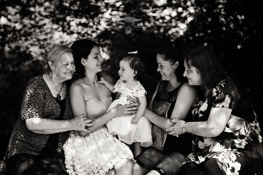 A black and white candid photograph of a multigenerational gathering outdoors with a small child in white at the center.