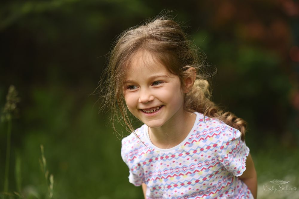 Child wearing a floral patterned dress smiles brightly while playing outdoors in a natural setting.