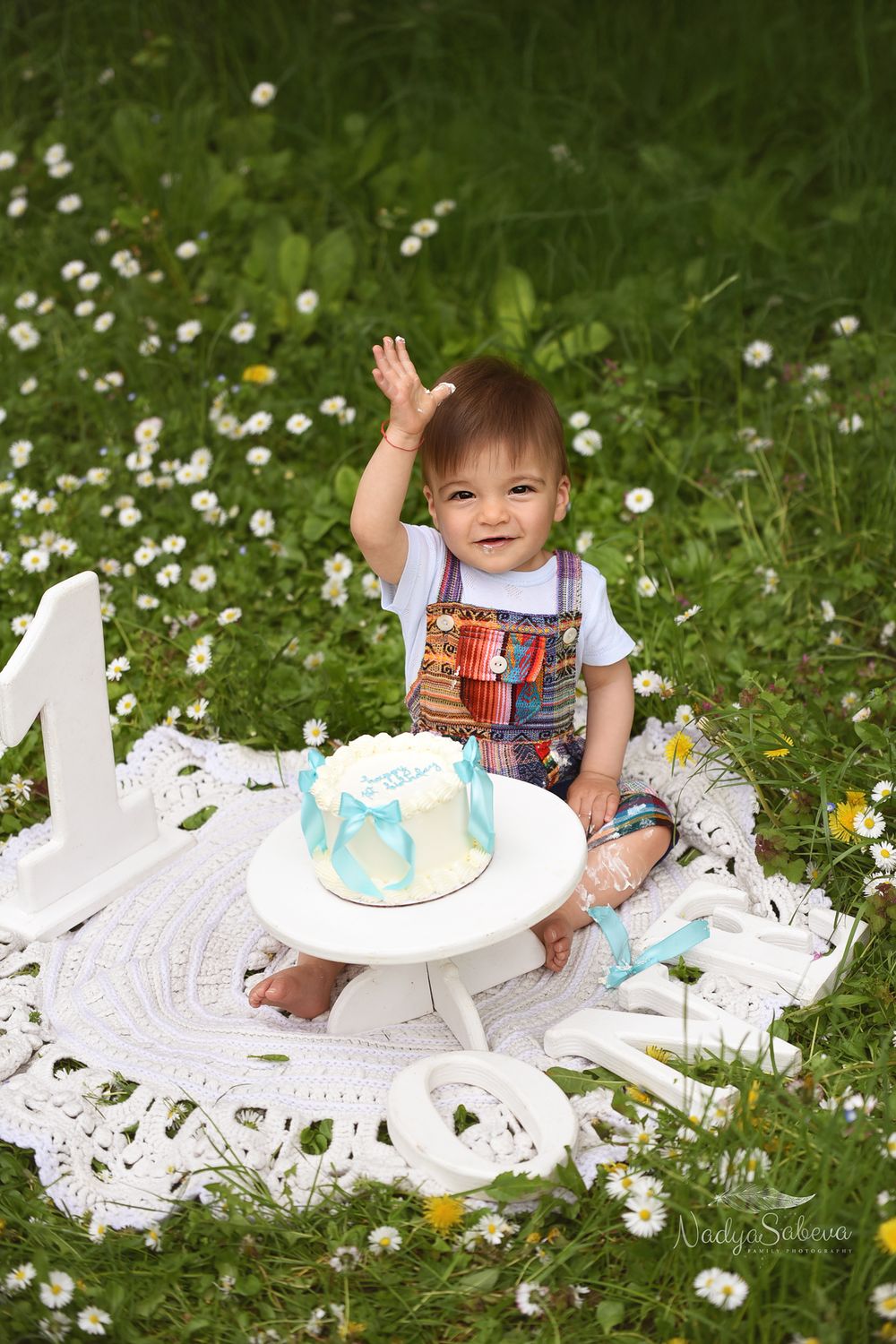 A baby in overalls sits on a white blanket in a flower-filled meadow with a blue and white cake for a first birthday celebration.