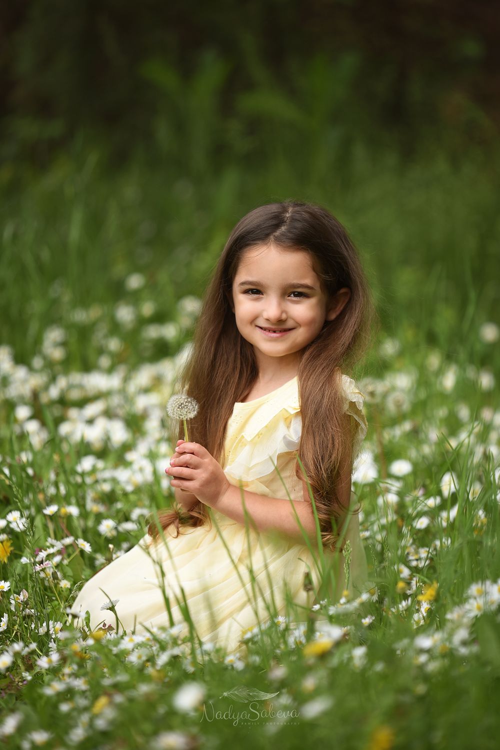A child in a yellow dress sits in a field of blooming white daisies while holding a dandelion flower on a summer day.