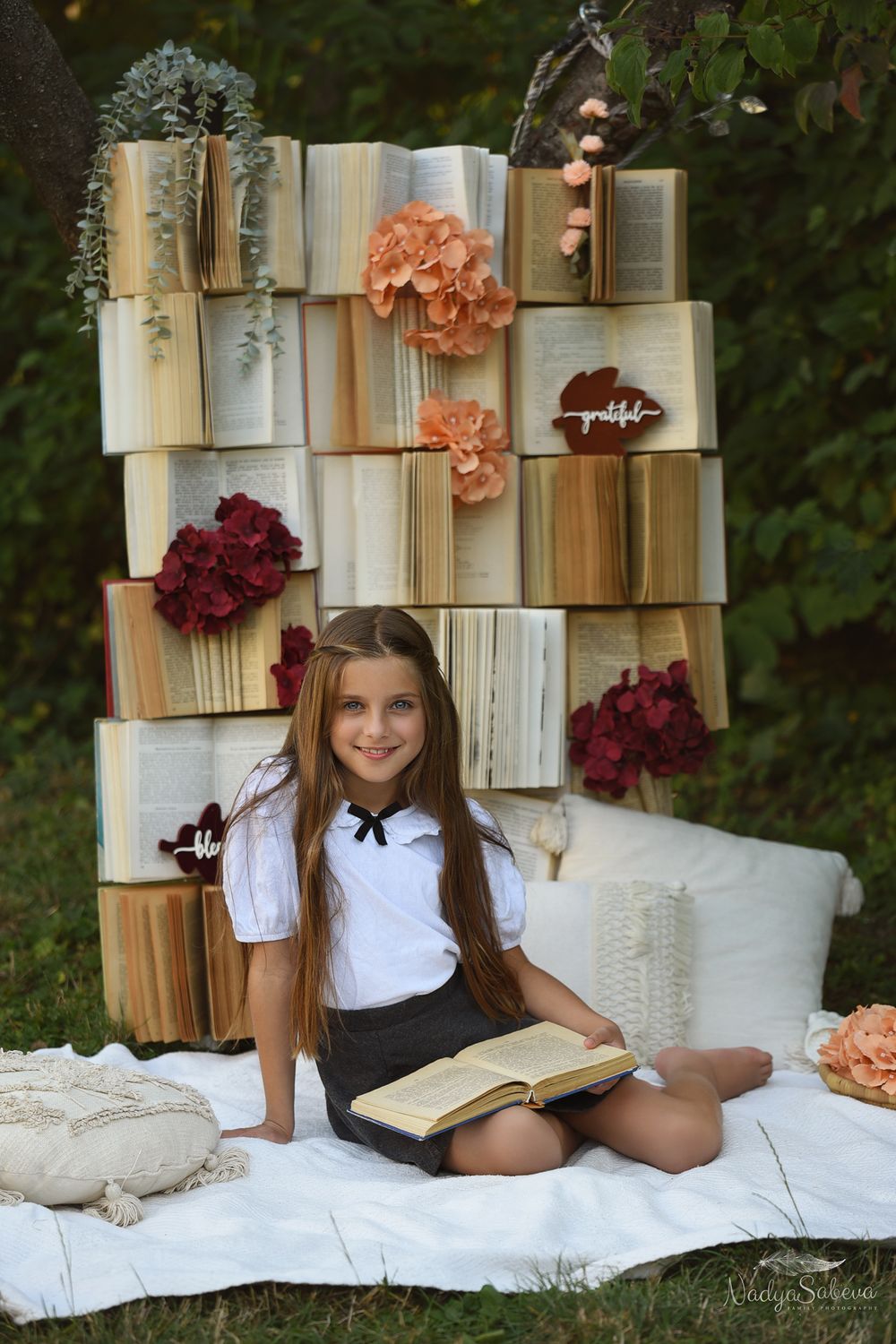 A girl reads a book on a white blanket against a backdrop of vintage books and pink flowers.