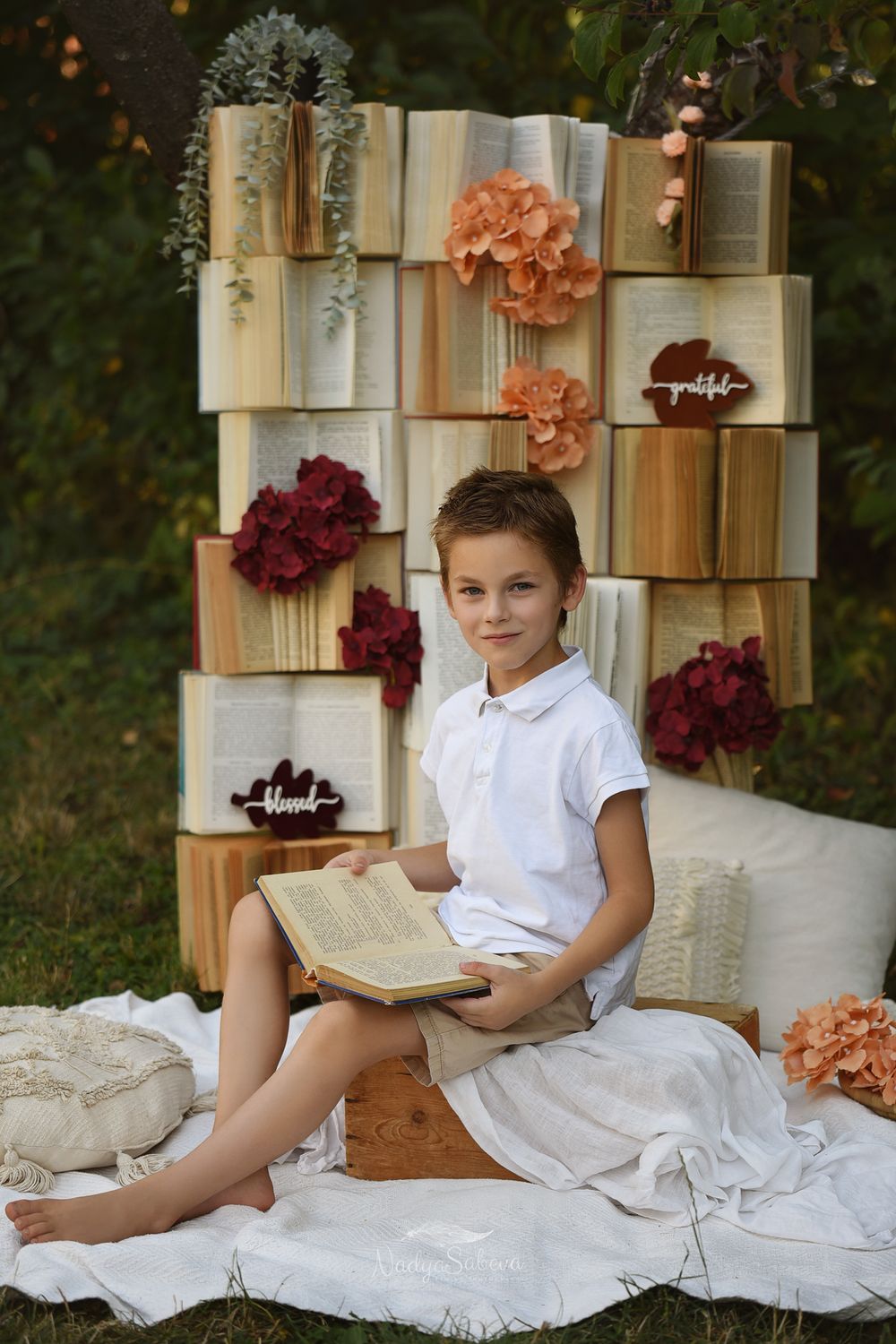 A child in white clothing reclines on white fabric among stacked vintage books and floral decor.