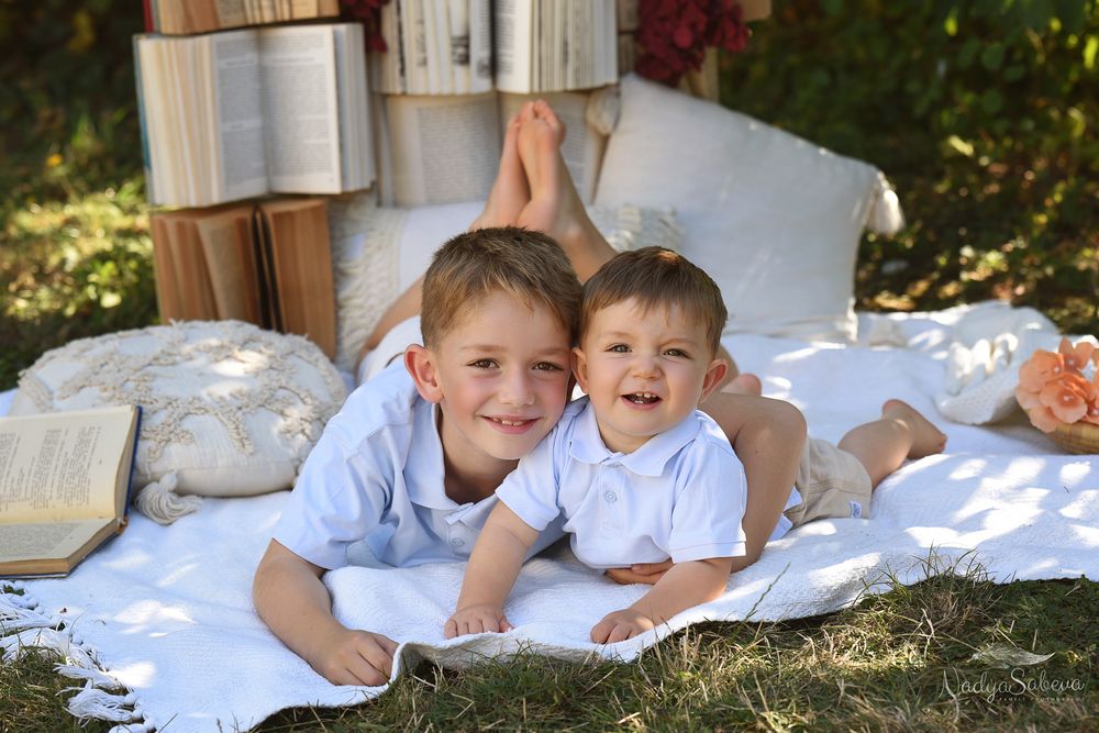 Children lie together on white bedding outdoors while reading books in the sunshine.