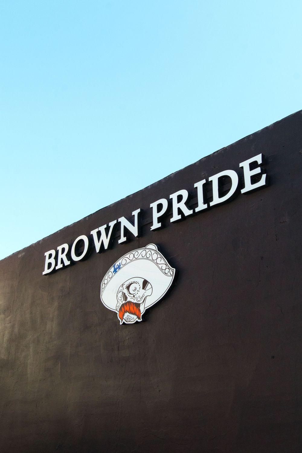 Brown Pride tattoo shop exterior wall sign with mariachi skull mascot against blue sky.