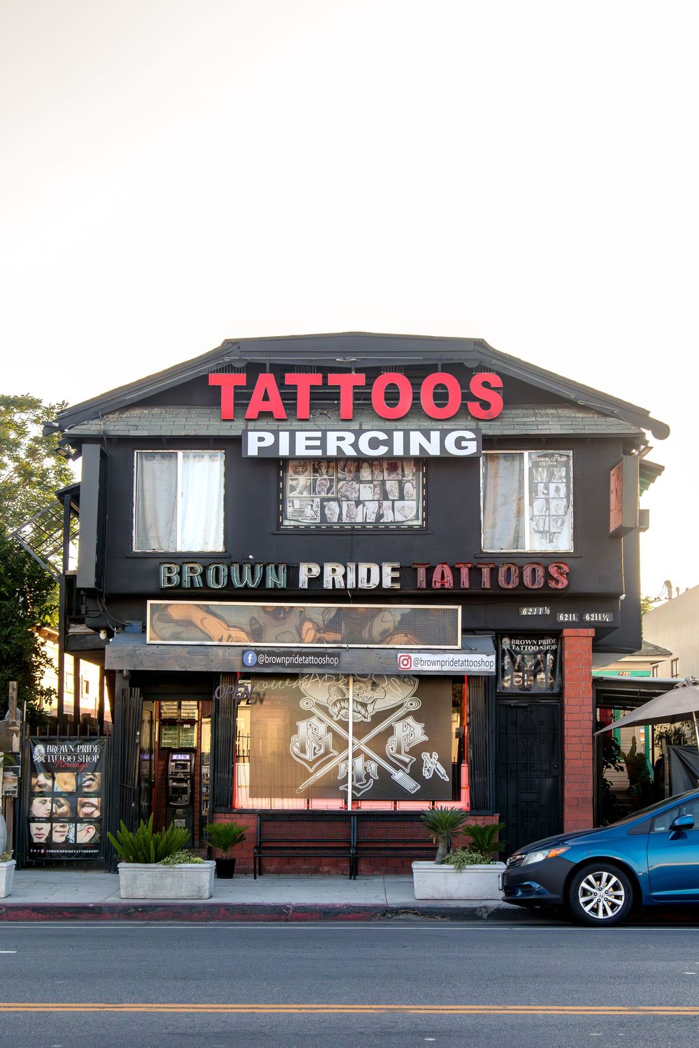 Brown Pride Tattoos and Piercing shop front view, black building with bold red signage at dusk.