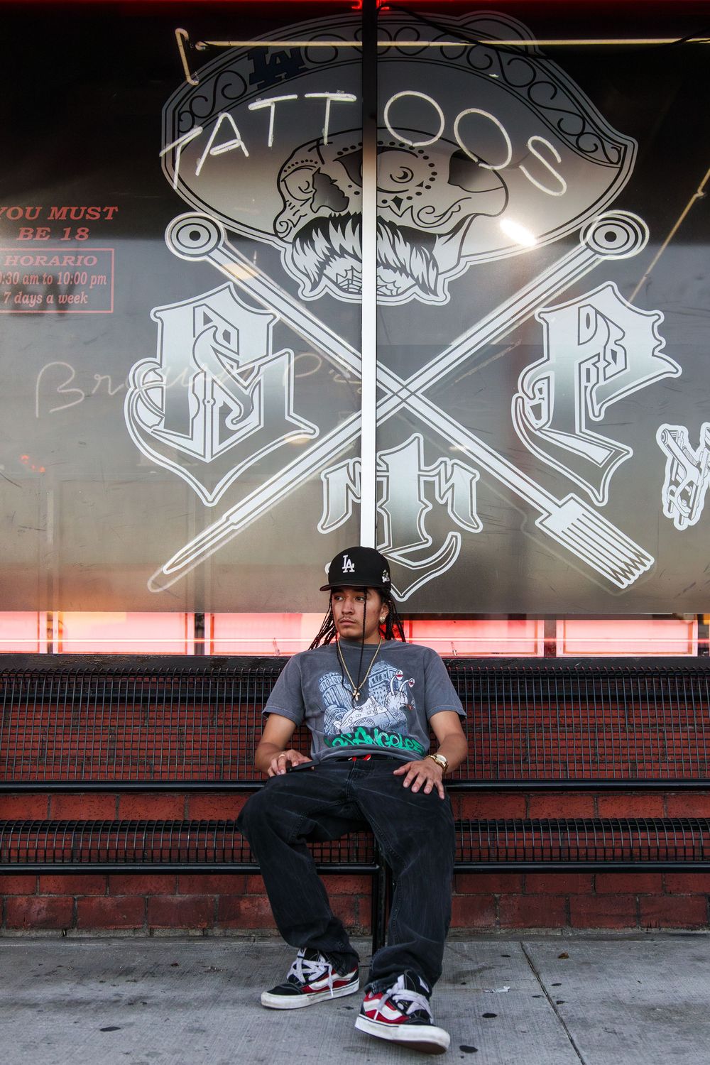 Tattoo artist with dreadlocks sitting outside Brown Pride Tattoos shop with neon window art.