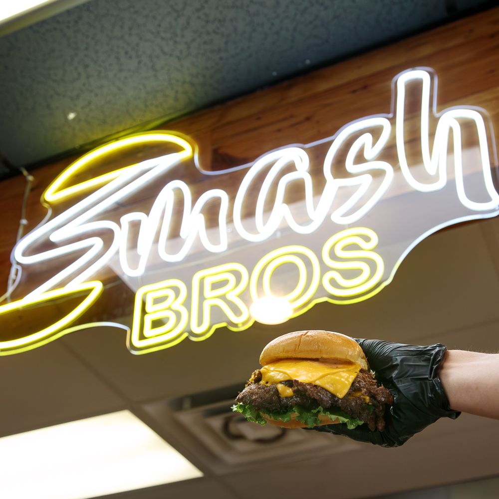 Gloved hand holding a smash burger under glowing Smash Bros neon sign.