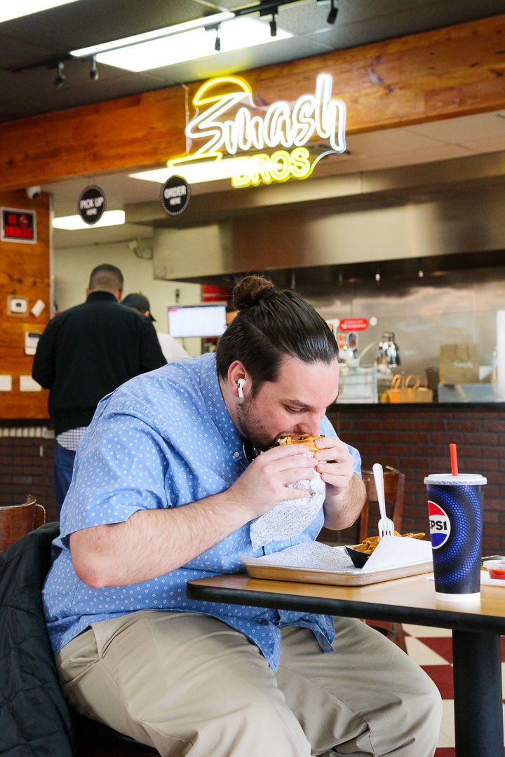 Man enthusiastically eating a smash burger inside Smash Bros restaurant with Pepsi drink.