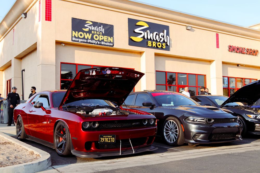 Two custom Dodge Challengers parked outside a Smash Bros Now Open restaurant location.