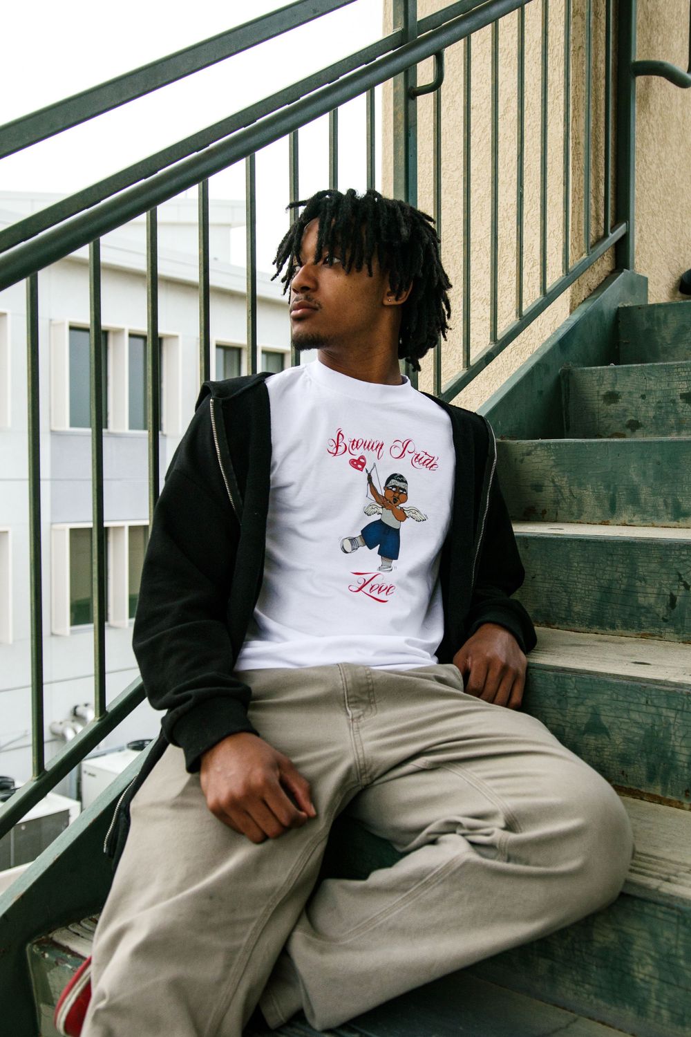 Man in white Brown Pride graphic tee sitting on outdoor stairs in casual streetwear.