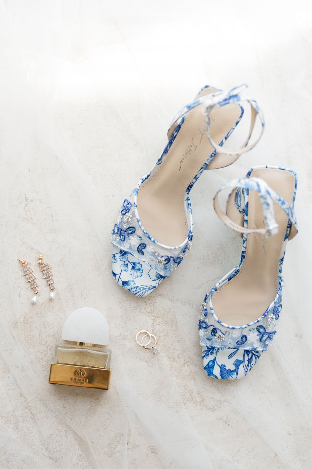 Blue floral bridal heels with ankle straps, gold drop earrings, perfume bottle, and wedding rings on marble surface.