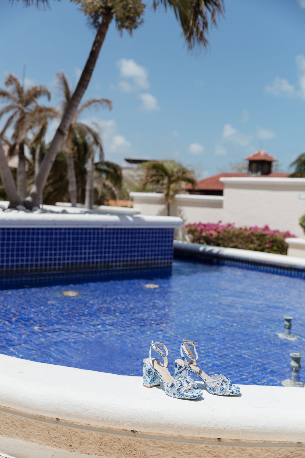 Blue floral heeled sandals placed on the edge of a tropical resort pool with palm trees and blue sky.