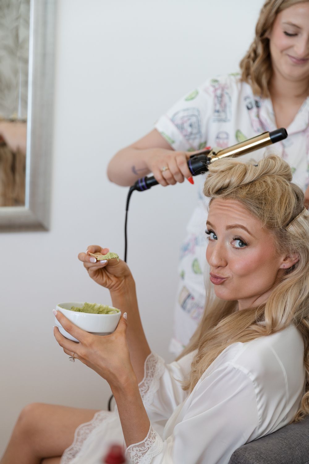 Blonde bride in white robe eating grapes while hairstylist curls her hair during wedding preparations.