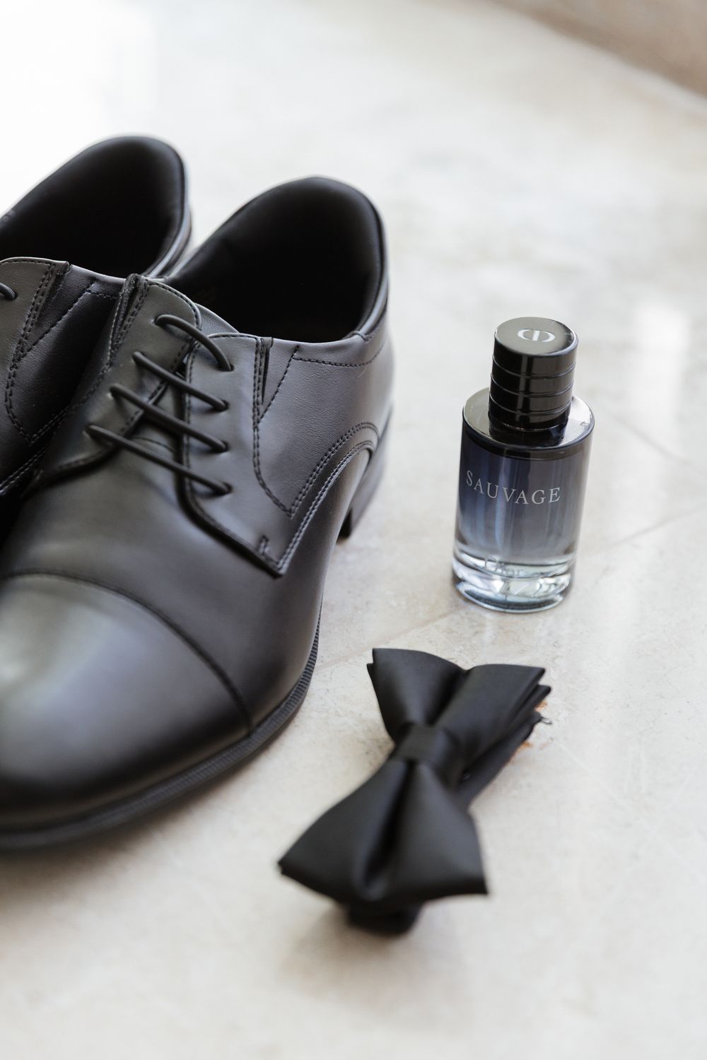 Black leather dress shoes, Dior Sauvage cologne bottle, and black bow tie arranged on marble surface for wedding day.
